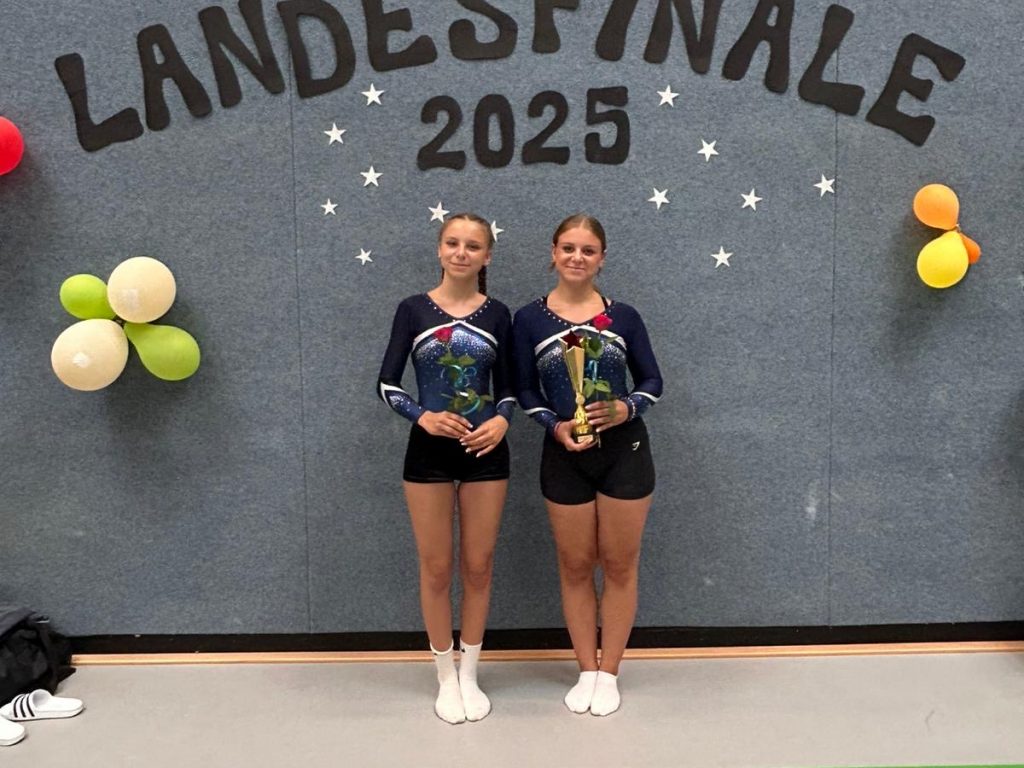 Zwei Turnerinnen stehen mit je einer roten Rose vor einer grauen Wand. Eine hält zusätzlich einen Pokal. Über ihnen ist der Schriftzug "Landesfinale 2025" auf die Wand geklebt.