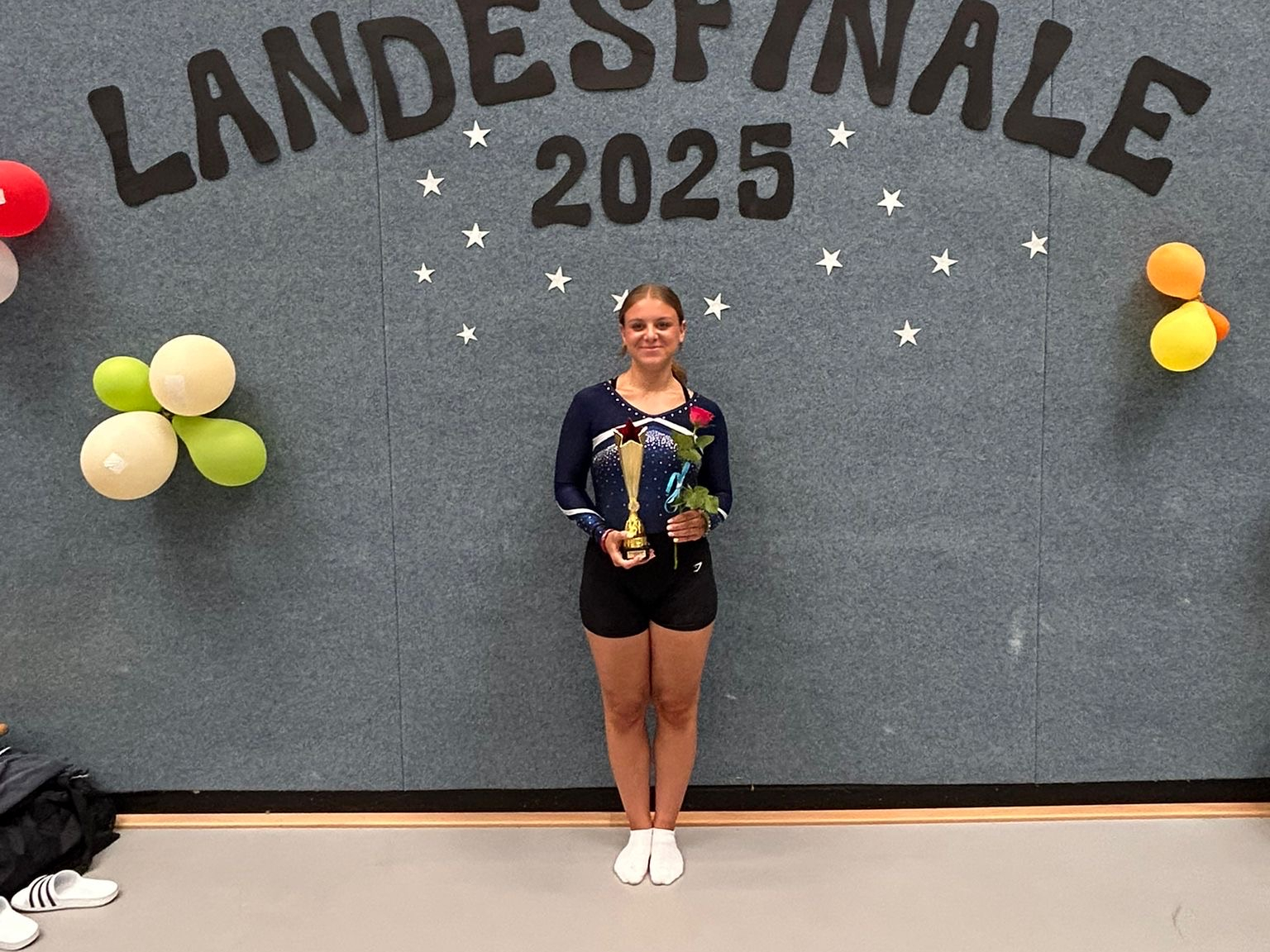Eine Turnerin steht mit einem Pokal und einer roten Rose vor einer grauen Wand. Über ihr ist der Schriftzug "Landesfinale 2025" auf die Wand geklebt.