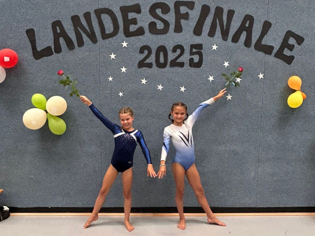 Zwei Turnerinnen stehen mit je einer roten Rose vor einer grauen Wand. Über ihnen ist der Schriftzug "Landesfinale 2025" auf die Wand geklebt.