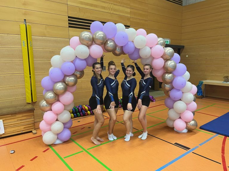 Vier Turnerinnen stehen unter einem Luftballonbogen aus weißen, rosanen, silbernen und lilanen Ballons.