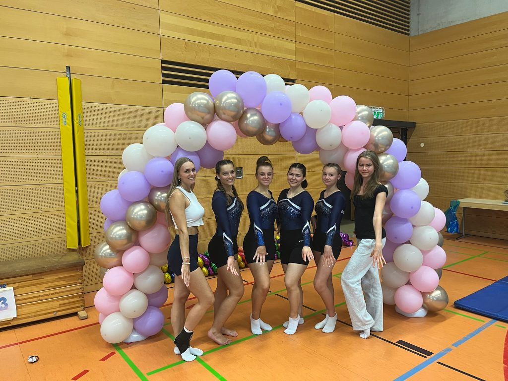 Vier Turnerinnen und zwei Betreuerinnen stehen unter einem Luftballonbogen aus weißen, rosanen, silbernen und lilanen Ballons.