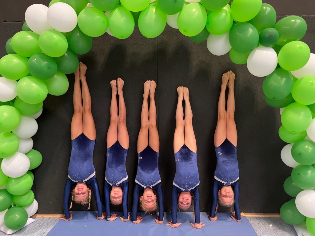 Fünf Turnerinnen machen Handstand unter einem Luftballonbogen aus grünen und weißen Ballons.