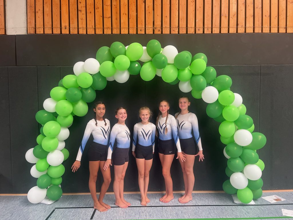 Fünf Turnerinnen stehen unter einem Luftballonbogen aus grünen und weißen Ballons.