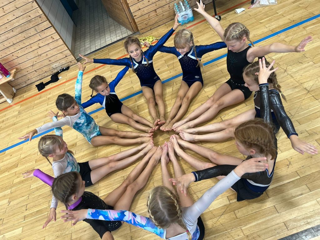 Zehn Turnerinnen sitzen im Langsitz mit gestreckten Armen im Kreis auf dem Boden.