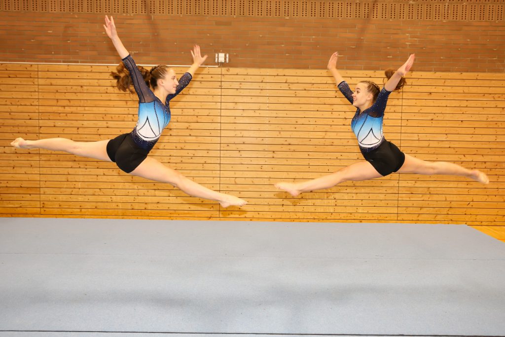 Bild von der Seite. Zwei Turnerinnen springen einen Spagatsprung aufeinander zu.