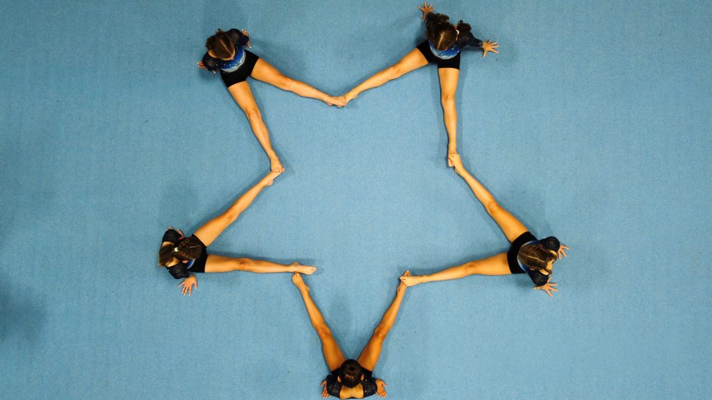 Gruppenbild von oben. Fünf Turnerinnen sitzen auf der blauen Bodenfläche. Mit ihren leicht gegrätschten Beinen formen sie einen 5-zackigen Stern.