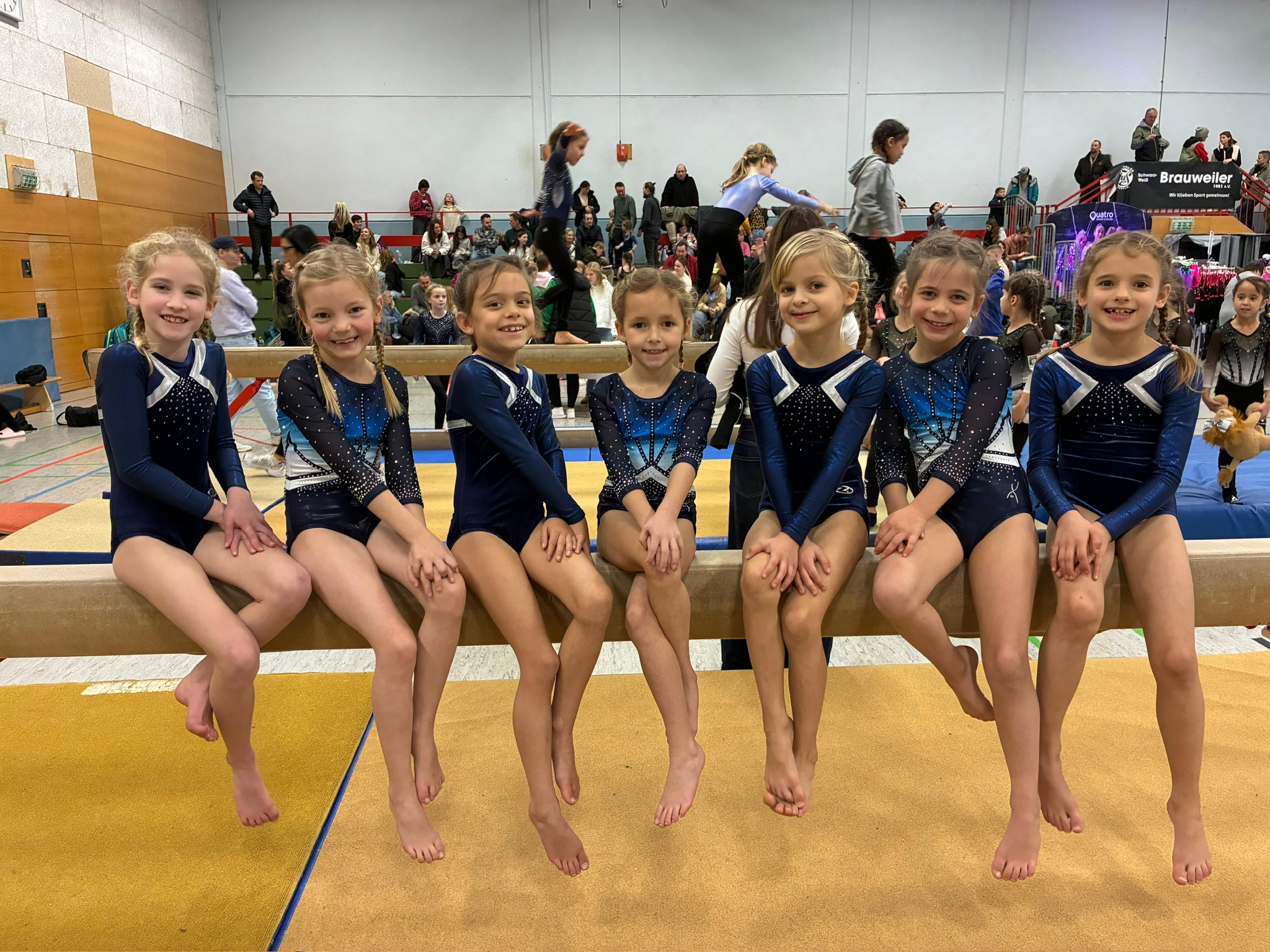 7 Turnerinnen sitzen nebeneinander auf dem Wschwebebalken und lächeln in die Kamera.