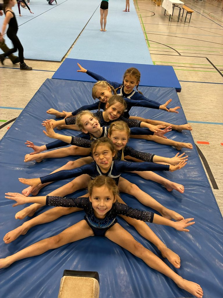 7 Turnerinnen sitzen hintereinander im Grätschsitz auf einer blauen Weichbodenmatte. Sie lachen und schneiden Grimassen.