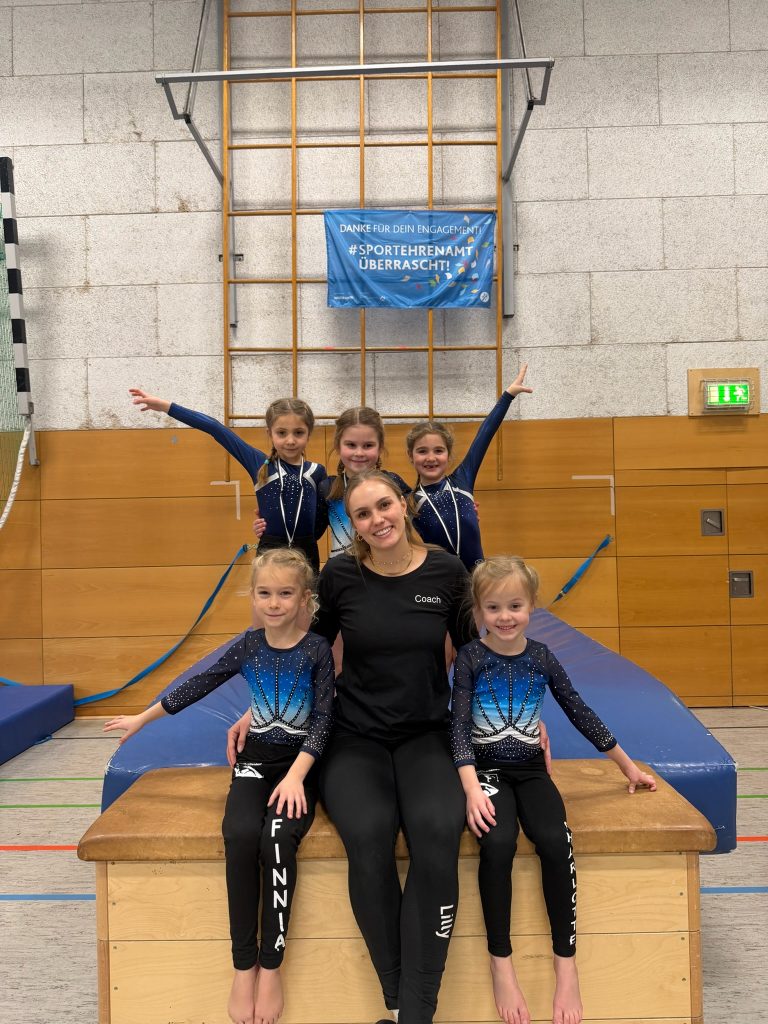 Zwei Turnerinnen sitzen links und rechts neben ihrer Trainerin auf einem Turnkasten. Hinter ihnen knien drei weitere Turnerinnen.