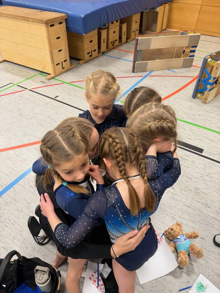 6 Turnerinnen stehen dicht gedrängt zusammen und umarmen sich gegenseitig. Blick von schräg oben auf ihre Köpfe.