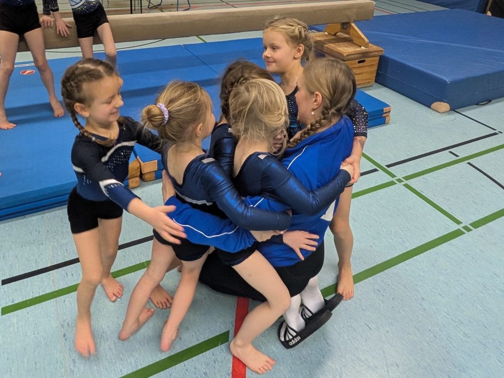 5 Turnerinnen umarmen ihre auf dem Boden kniende Trainerin.