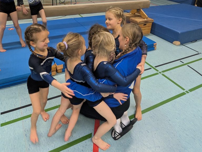 5 Turnerinnen umarmen ihre auf dem Boden kniende Trainerin.