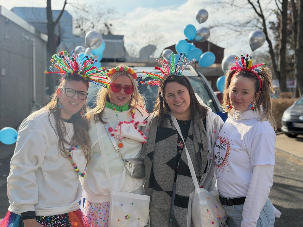 Gruppenfoto: Vier Teilnehmenden in bunten Konfetti-Kostümen vor einem mit blauen und silbernen Luftballons geschmückten Auto.