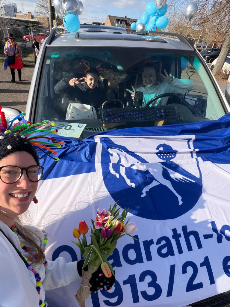 Gruppenfoto: Eine Teilnehmende in buntem Konfetti-Kostüm vor einem mit Vereinsfahne und blauen und silbernen Luftballons geschmückten Auto. Im Auto zwei weitere Teilnehmende, die Grimassen schneiden.