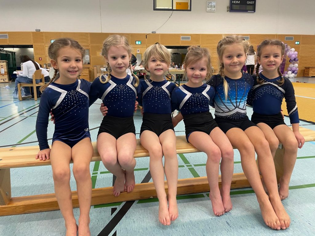 6 Turnerinnen sitzen nebeneinander auf einer Turnbank und lächeln in die Kamera.