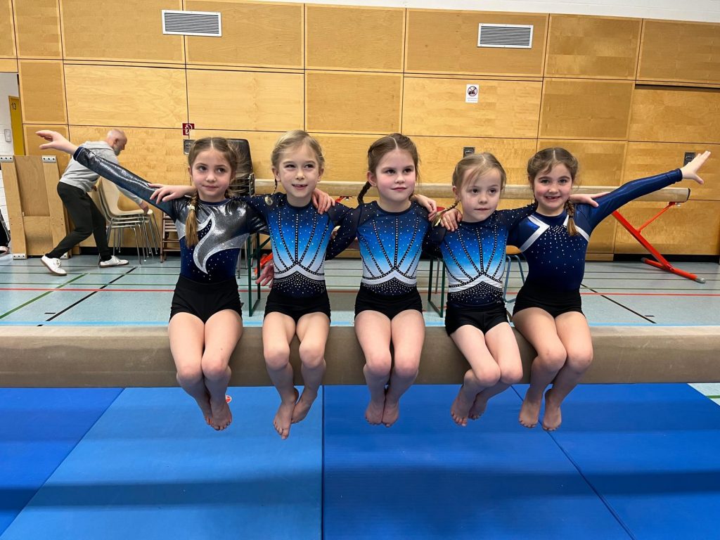 5 Turnerinnen sitzen nebeneinander auf einem Schwebebalken und lächeln in die Kamera.