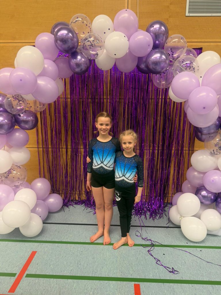 2 Turnerinnen stehen unter einem Luftballonbogen und lächeln in die Kamera.