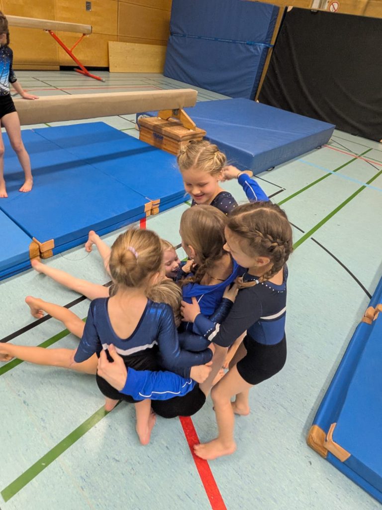 5 Turnerinnen umarmen ihre auf dem Boden sitzende Trainerin.