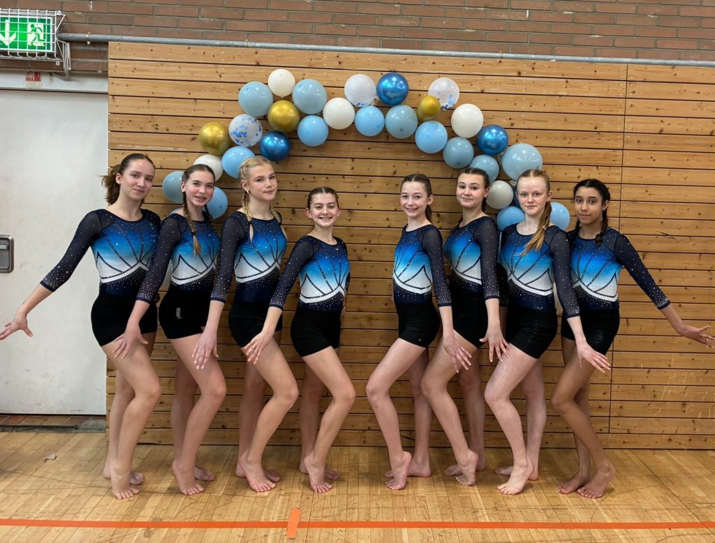 8 Turnerinnen stehen vor einem Luftballonbogen und lächeln in die Kamera.