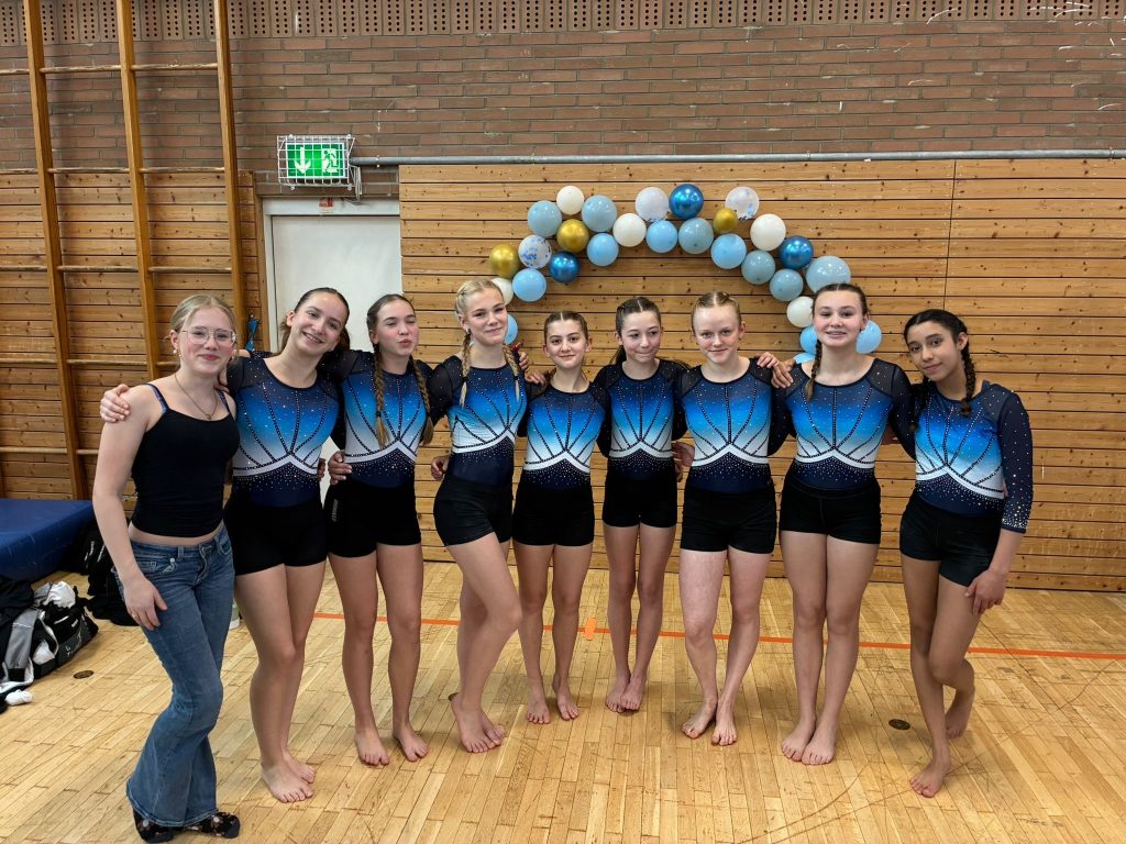8 Turnerinnen und eine weiteres Mädchen stehen vor einem Luftballonbogen und lächeln in die Kamera.