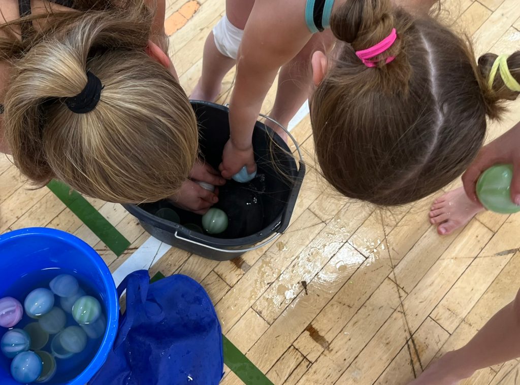 Blick von oben auf die Köpfe zweier Mädels, die Wasserbomben in einem Eimer befüllen. Neben ihnen steht ein Eimer mit weiteren befüllten Wasserbomben.