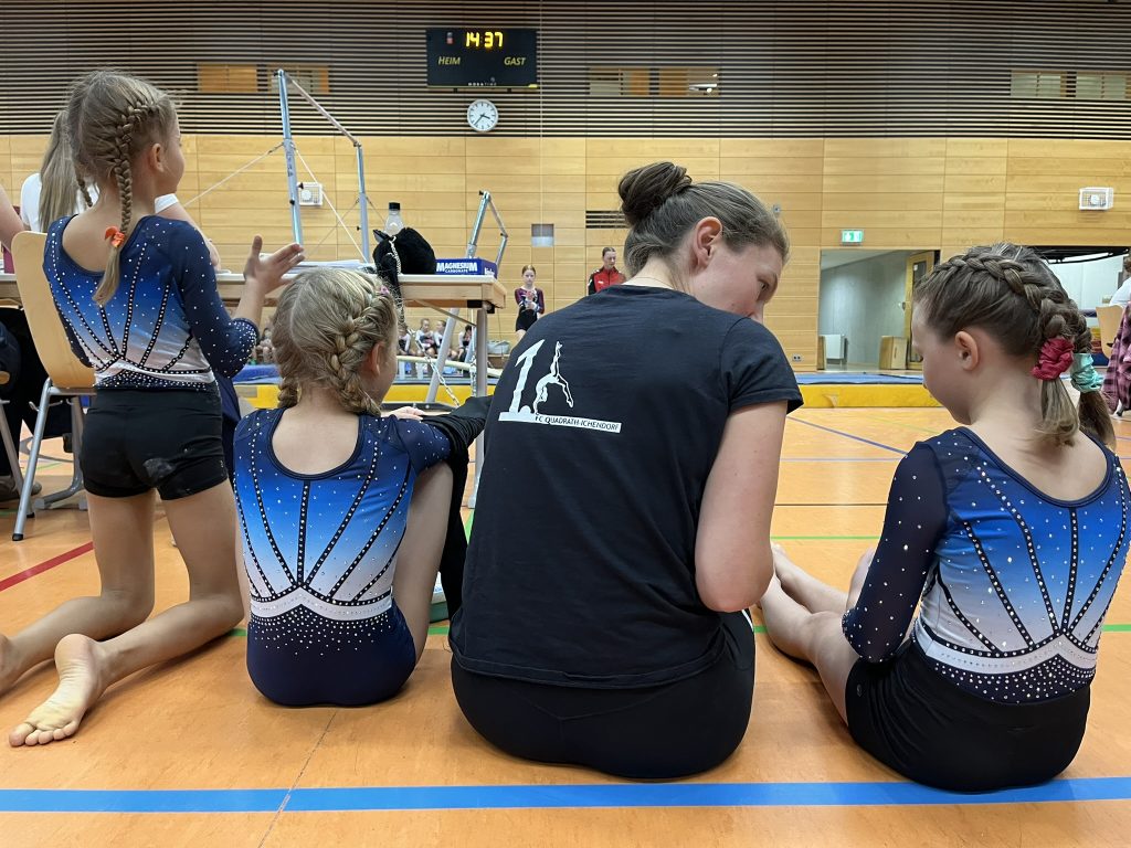 Eine Trainerin und drei Turnerinnen von hinten. Sie sitzen auf dem Boden. Die Trainerin spricht mit der Turnerin rechts neben ihr.