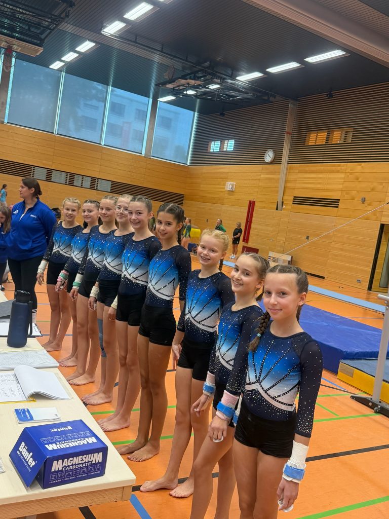 Neun Turnerinnen in einheitlichen blauen Wettkampfanzügen stehen nebeneinander vor dem Tisch des Kampfgerichtes. Links neben ihnen steht eine Trainerin.