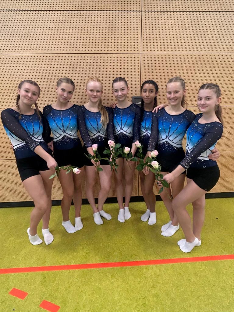 Sieben Turnerinnen in einheitlichen blauen Wettkampfanzügen stehen nebeneinander vor einer Holzprallwand. Sie halten Rosen in den Händen.